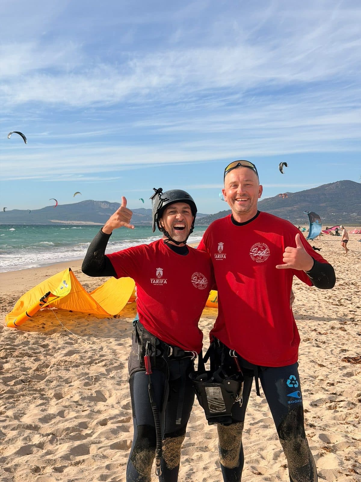 Zwei Kiter in roten Shirts zeigen am Strand das Shaka-Zeichen